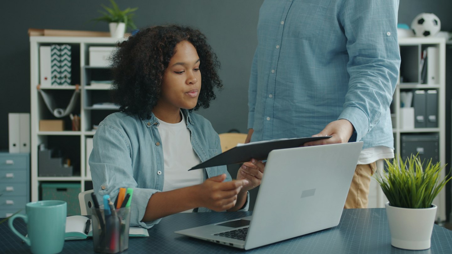 Zwei Kollegen arbeiten an einem Schreibtisch mit Laptop und Ordner zusammen.